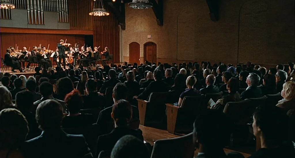 Chloe (2009) movie still: wide — A dimly lit, large indoor auditorium features rows of audience members facing a wooden stage. On the stage, a…