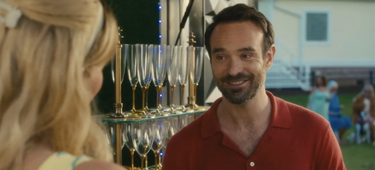 Merv (2025) movie still: medium — A sunny backyard party features a glass shelving unit filled with clear champagne flutes. A man in a terracotta…