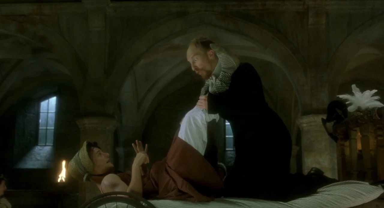 Quills (2000) movie still: medium — A stone vaulted cellar features rough masonry walls and a small fire burning below. A man in a dark coat reaches…