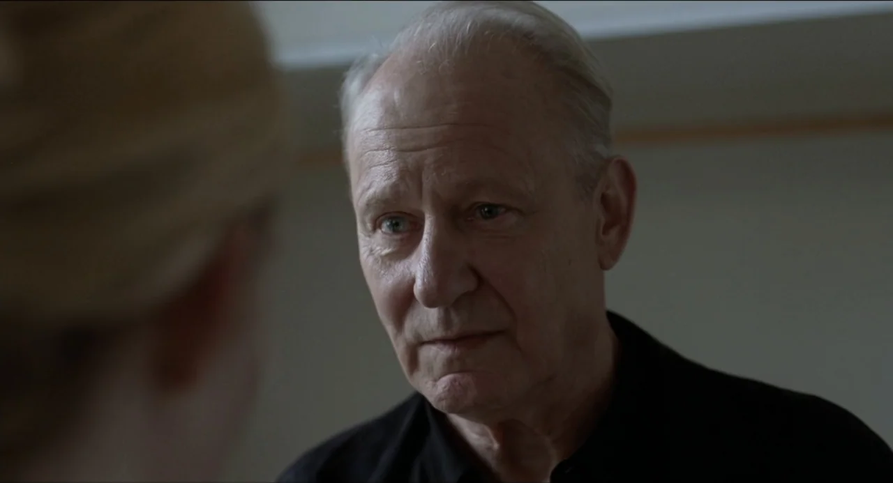 Sentimental Value (2025) movie still: closeup — An interior space with a neutral wall in the background. An older man is viewed over the shoulder of…