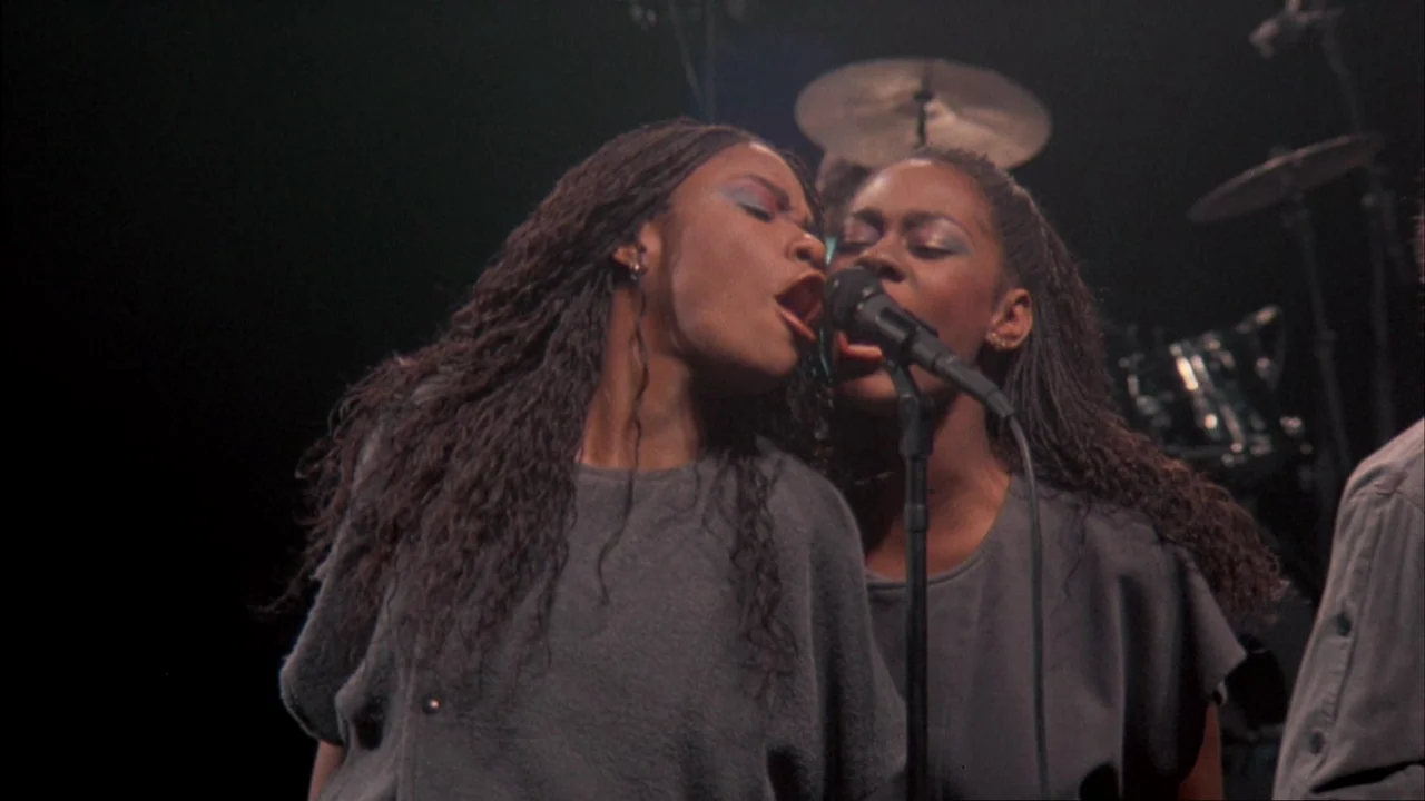 Stop Making Sense (1984) movie still: medium — A dark, dimly lit studio space with silhouettes of percussion equipment visible behind singers. Two…