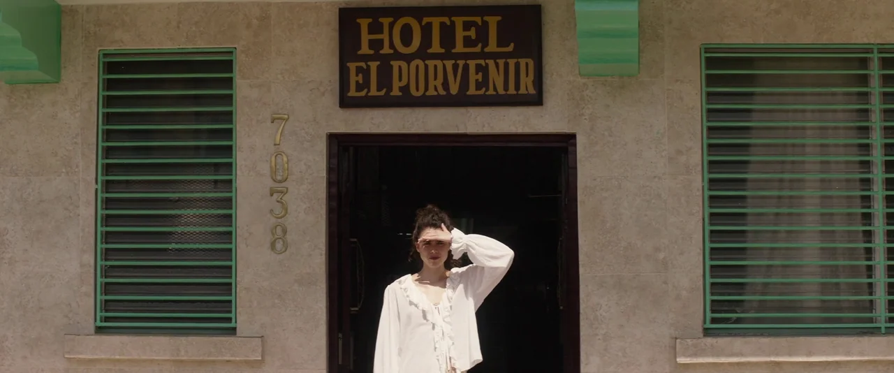 Stars At Noon (2022) movie still: medium — A stone building exterior with green metal window shutters and a hotel signage board. A woman stands in…