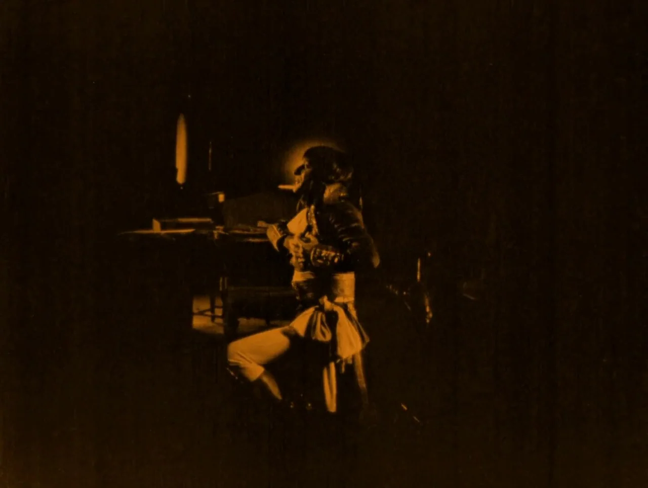 Napoleon (1927) movie still: medium — A man in 18th-century attire sits in a darkened room with furniture nearby. The man is reclined on a wooden…
