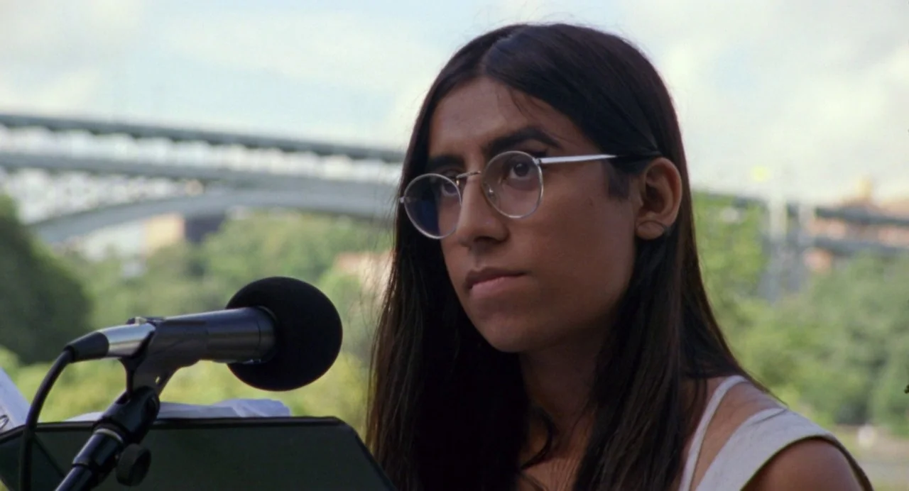 So Pretty (2019) movie still: closeup — An outdoor park setting features a person standing at a microphone before a bridge. The person looks toward…