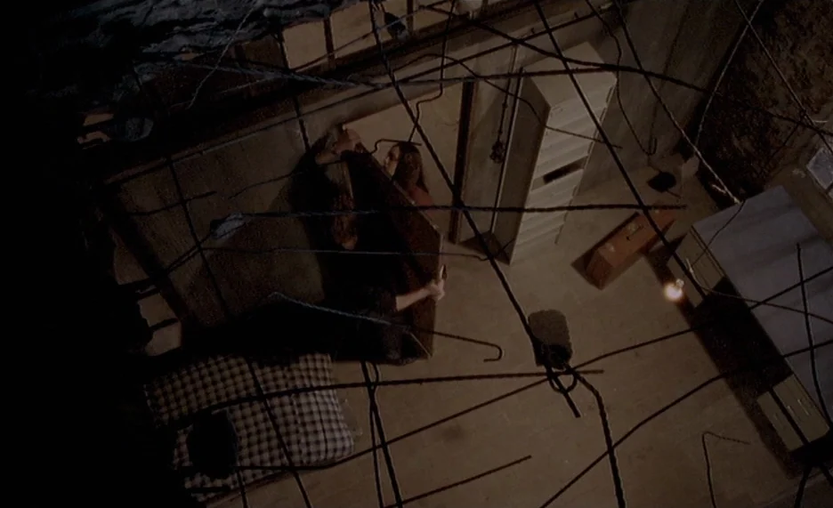Pola X (1999) movie still: wide — A cramped, dimly lit room filled with a tangled web of dark wires overhead. A man and woman hold a large piece of…