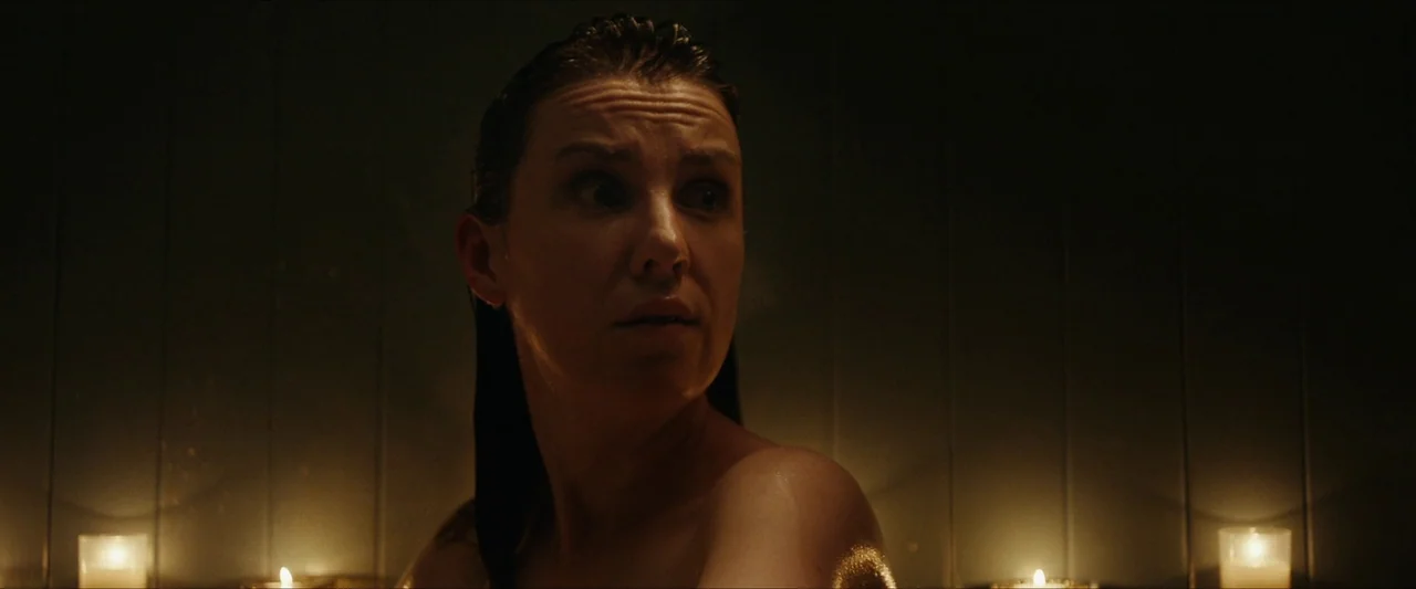 Fréwaka (2024) movie still: closeup — A dimly lit room with vertical wood paneled walls in the background. A woman with wet hair looks toward the…