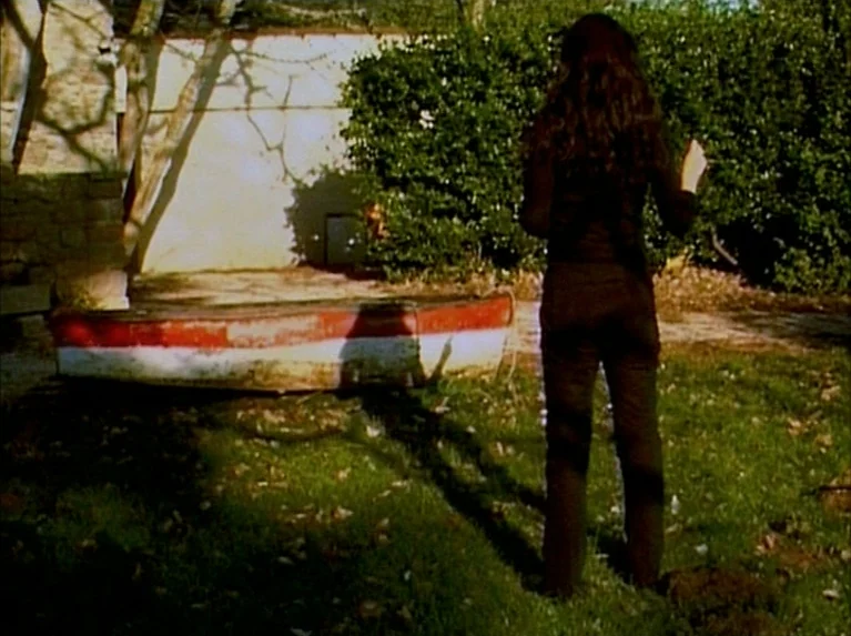 Eloge de L’amour (2001) movie still: fullbody — A sunlit backyard features a small abandoned rowboat resting on a patch of grass. A woman stands…