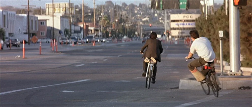 I Heart Huckabees (2004) movie still: wide — A wide, empty city street during the day with commercial buildings and traffic lights. Two men cycle…