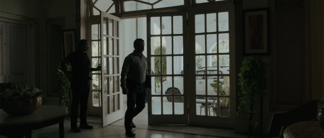 Nishaanchi 2 (2025) movie still: fullbody — A bright doorway leads from an outdoor area into a dimly lit building foyer. Two men walk through the…