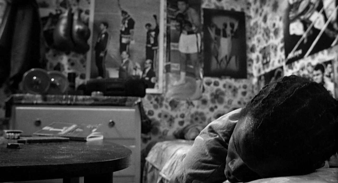 La Haine (1995) movie still: medium — A dark bedroom with floral wallpaper is adorned with multiple posters of boxers. A young man rests with his…