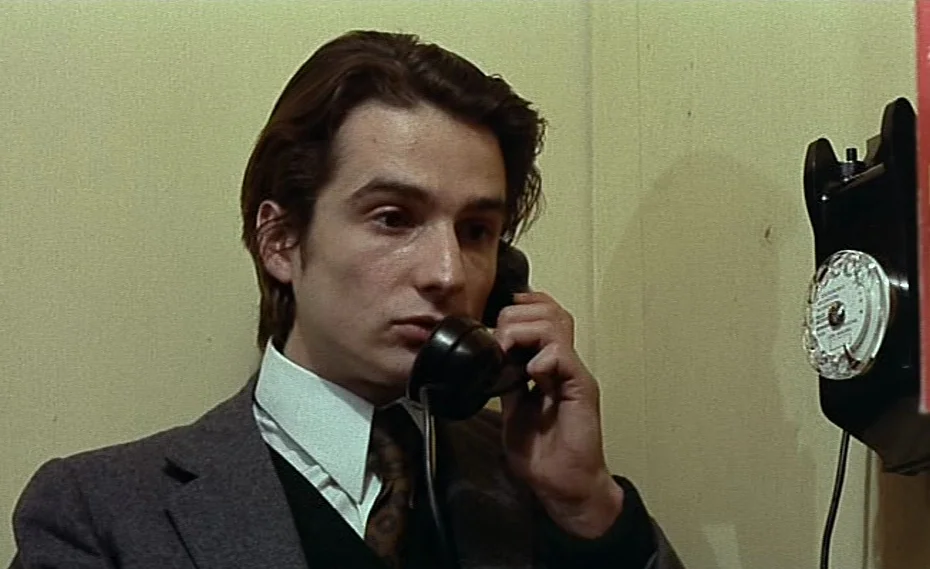 Bed & Board (1970) movie still: closeup — A small, plain interior with light beige walls and a mounted rotary telephone. A young man wearing a suit…
