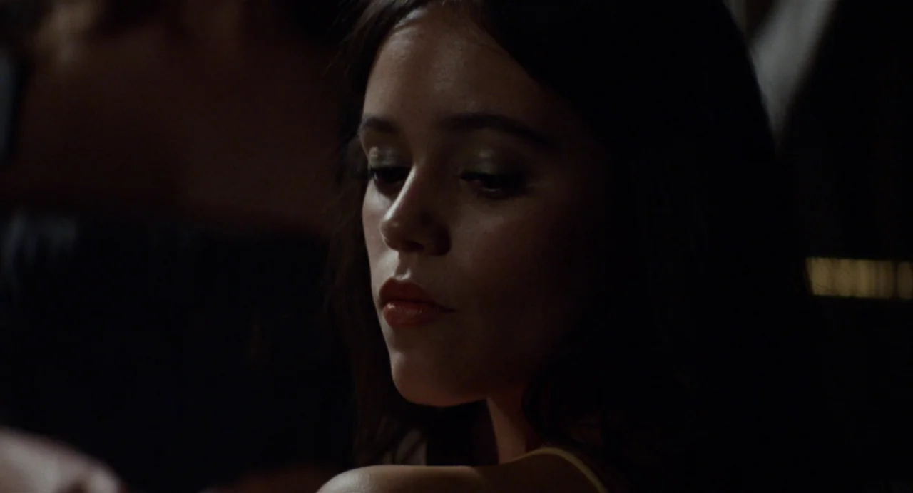 X (2022) movie still: closeup — A dimly lit indoor space with a dark, indistinct background. A young woman looks down with a neutral facial…