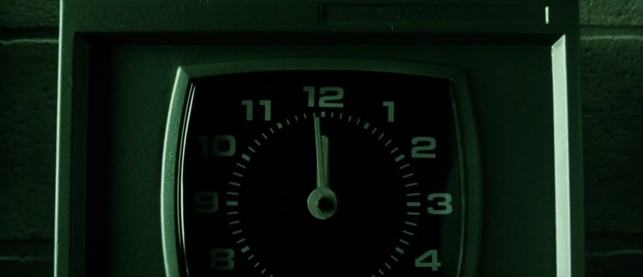 The Matrix Reloaded (2003) movie still: closeup — A close-up view of a dark analog wall clock mounted on a brick surface. The clock hands indicate…