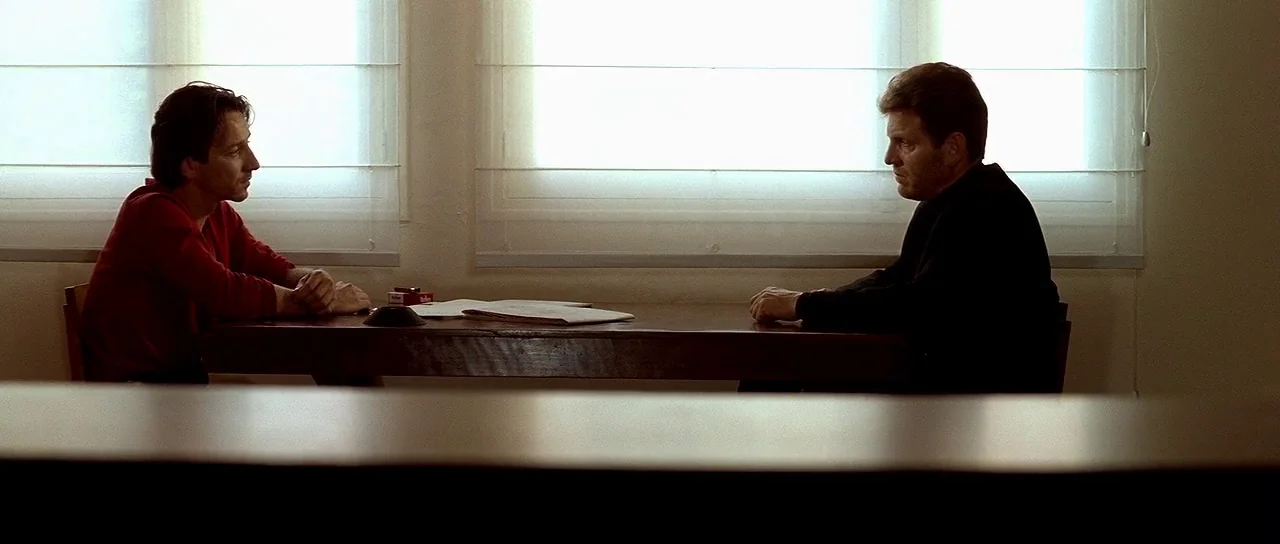 Nikita (1990) movie still: medium — A sparse interrogation room features white walls and two large windows with horizontal blinds. Two men sit at a…