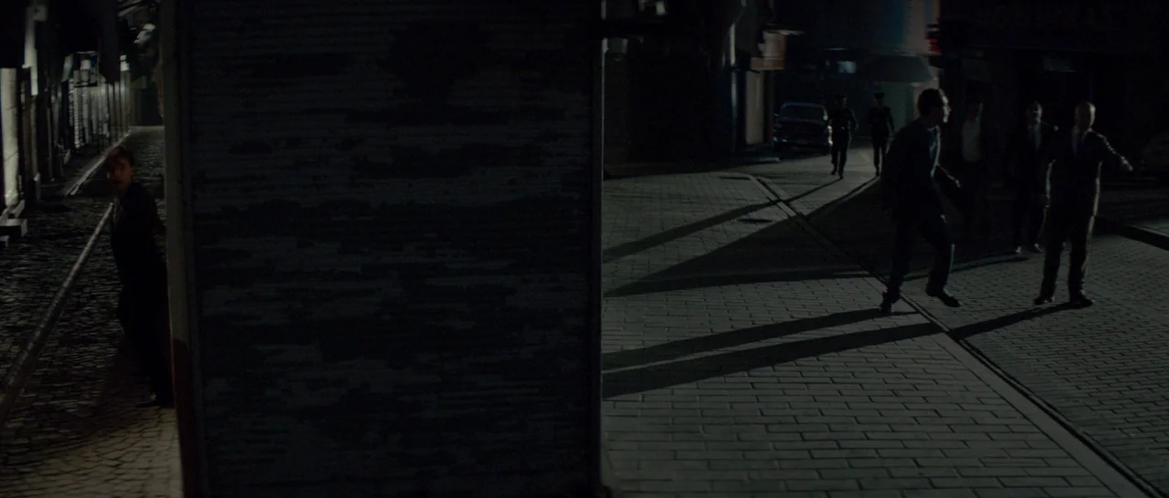 The Two Faces of January (2014) movie still: wide — A dark night plaza with cobblestone textures and sharp shadows from overhead lighting. One man…