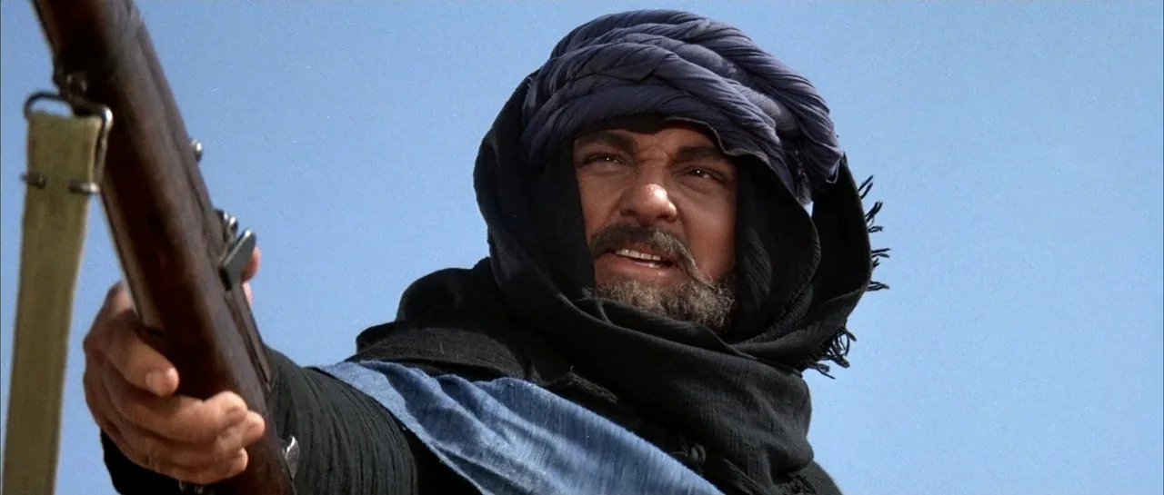 Sahara (1995) movie still: medium — An open desert environment under a clear blue sky. A man with a dark beard holds a rifle pointed forward.
