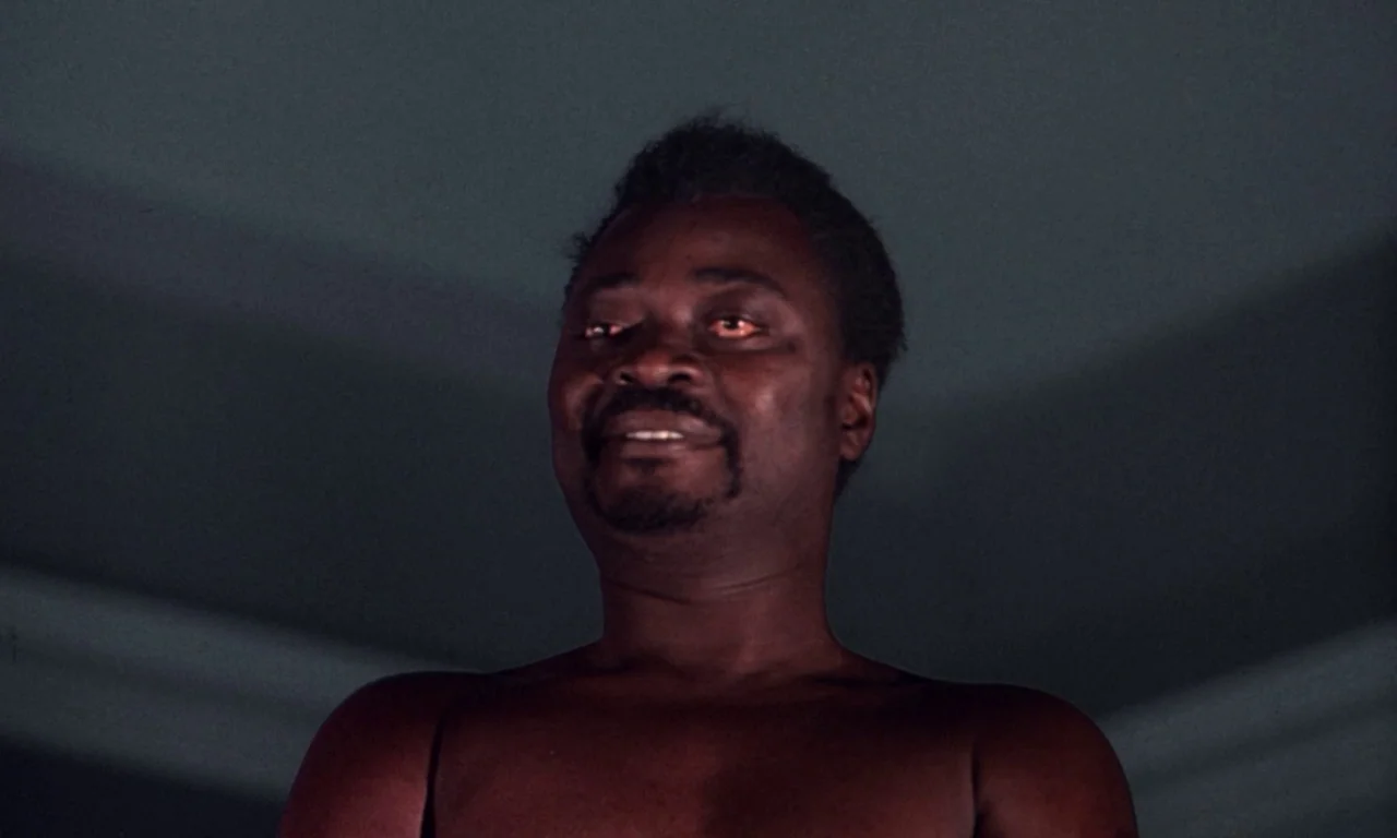 The Devil Rides Out (1968) movie still: closeup — An interior space with dark walls and subtle shadow contours. A shirtless man stares forward with…