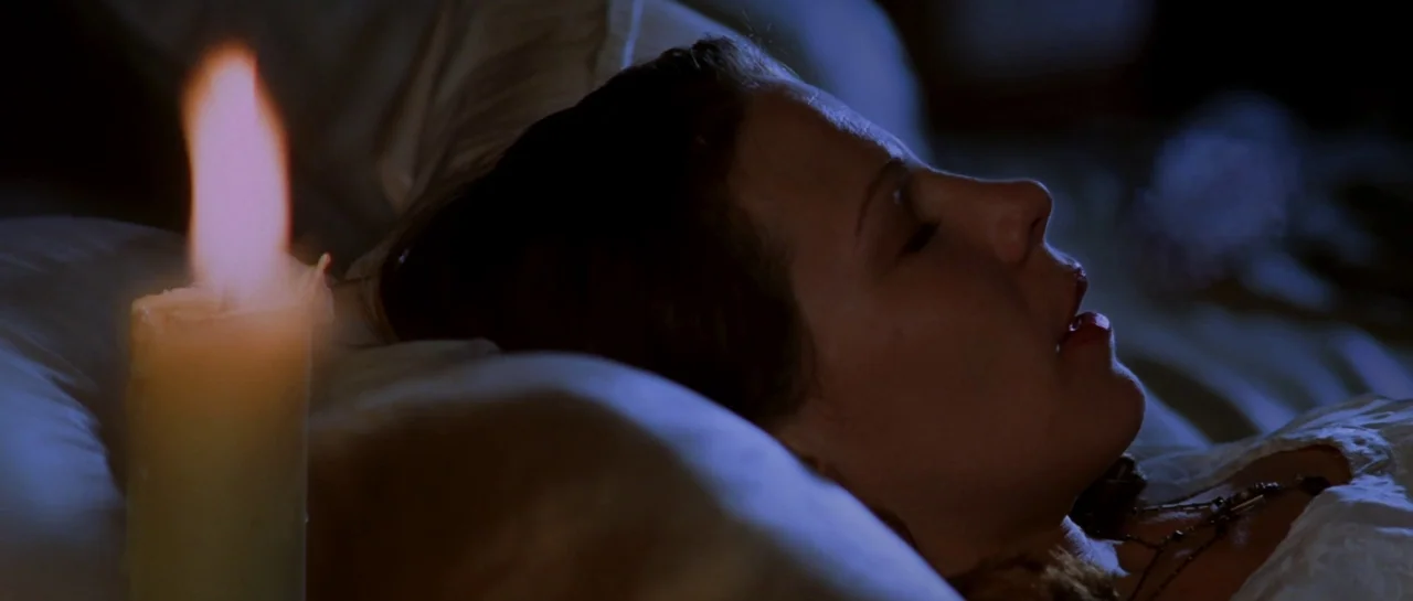 The Haunting (1999) movie still: closeup — A woman lies on white pillows in a dimly lit space during the night. A single burning candle sits in the…