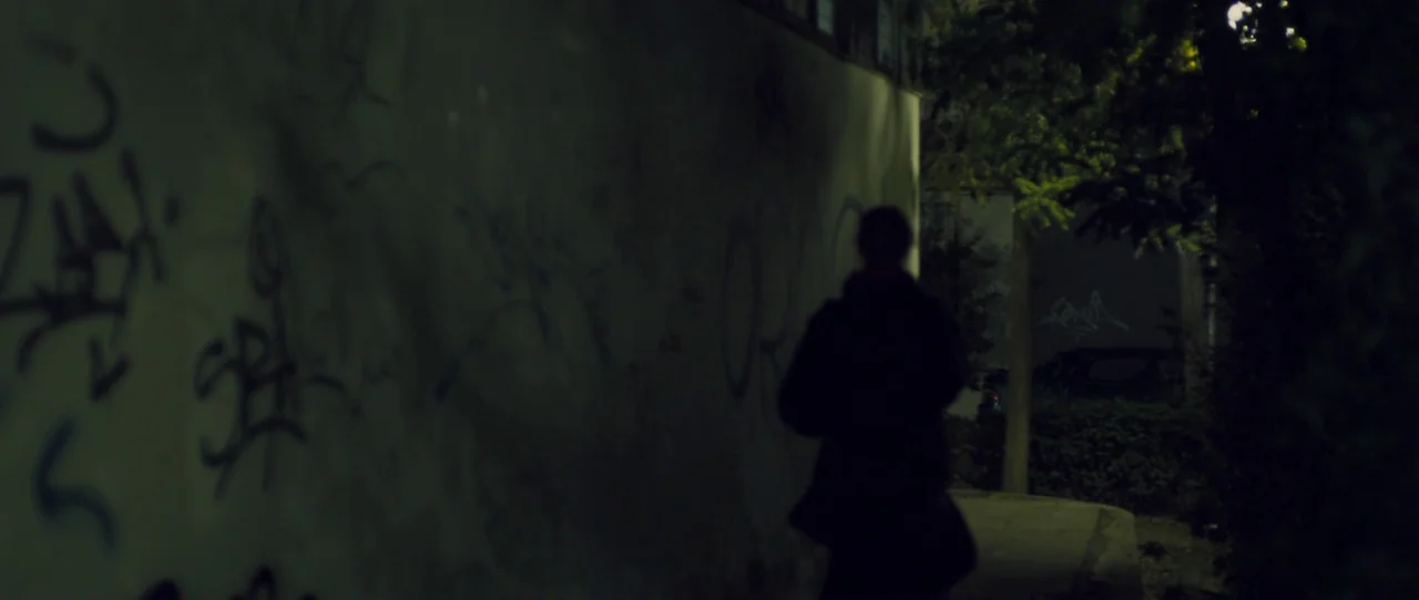 Alps (2011) movie still: medium — A dark urban sidewalk beside a tall concrete wall covered in black graffiti marks. A lone hooded figure walks away…