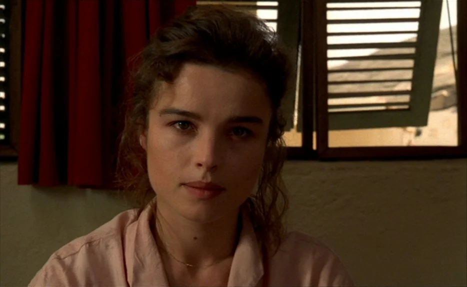 Chocolat (1988) movie still: closeup — A room features a window with horizontal slatted shutters and dark red curtains. The woman sits facing the…