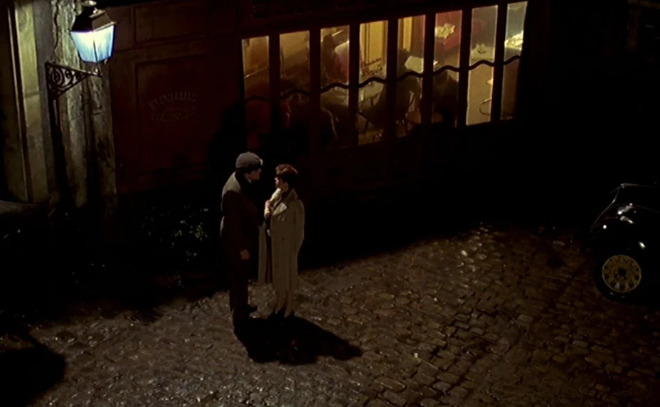The Last Metro (1980) movie still: wide — A damp, dark cobblestone street features an old building with glowing window panels. A man and a woman…