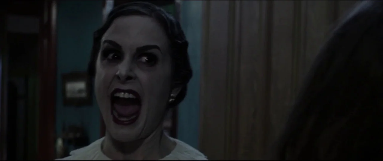 Insidious: Chapter 2 (2013) movie still: closeup — A dimly lit residential corridor features muted teal walls and dark wooden door frames. A woman…