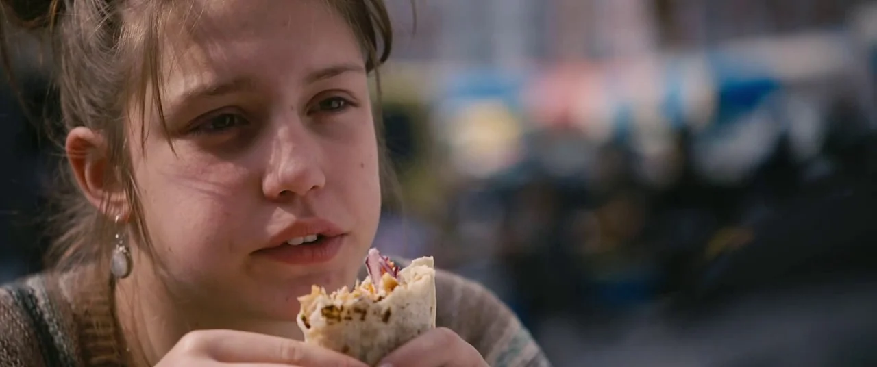 Blue Is The Warmest Colour (2013) movie still: closeup — An outdoor seating area features blurred urban activity in the background. A young woman…