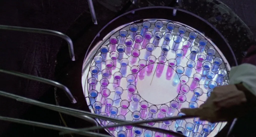 Demons of the Mind (1972) movie still: closeup — A circular array of glass test tubes is held in a metal rack structure. The tubes are backlit,…