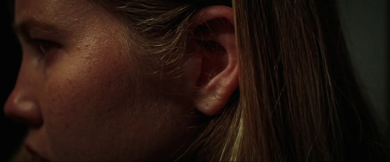 Cuckoo (2024) movie still: closeup — A close-up view focuses on the side profile of a woman's face and ear. The lighting highlights skin texture…