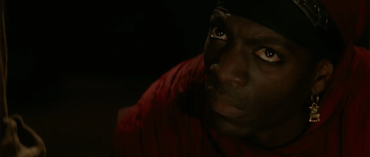 The Mummy Returns (2001) movie still: closeup — An interior space shrouded in deep darkness with minimal ambient light. A man in red clothing looks…