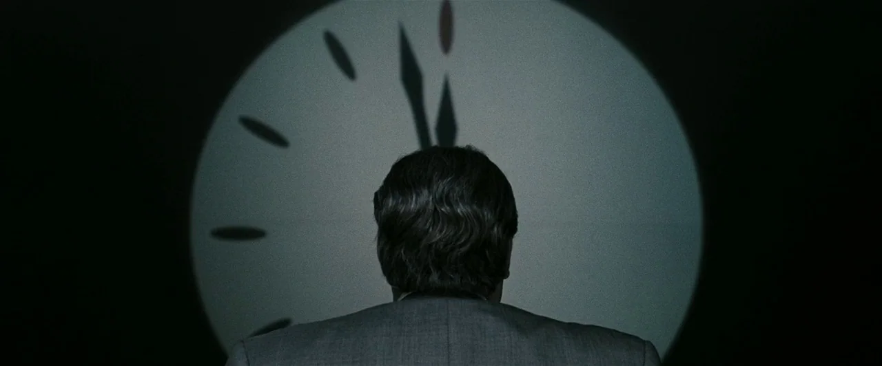 Watchmen (2009) movie still: medium — A man stands in a pitch black room facing a large circular projection. The projection displays a clock face…