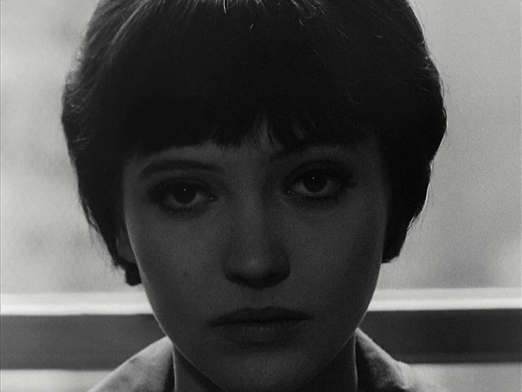 Vivre Sa Vie (1962) movie still: closeup — A dark indoor space with a horizontal light bar visible behind the subject. A young woman with short dark…