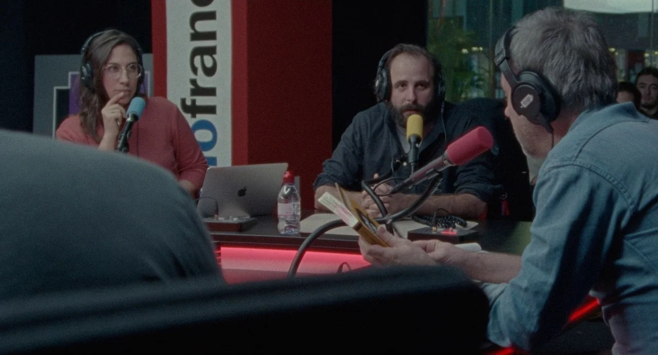 Non-Fiction (2018) movie still: medium — A modern radio broadcast booth features sound-dampening panels and branding on a red pillar. Three…