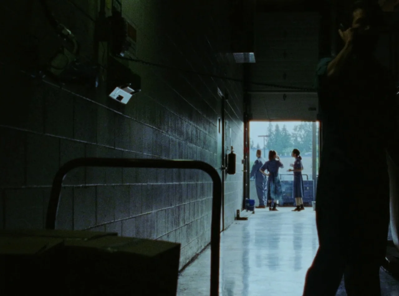 Riceboy Sleeps (2022) movie still: wide — A dimly lit concrete corridor leads to a brightly lit open loading dock area. Three figures stand in the…