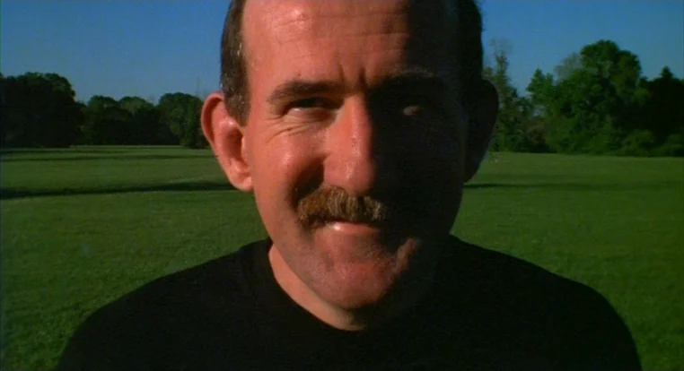 Schizopolis movie still: closeup — An open grassy field under a bright clear sky during the day. A man faces forward while positioned in the center…