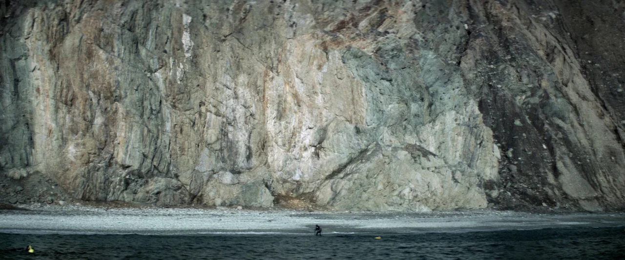 Chevalier (2015) movie still: wide — A massive, craggy rock cliff rises vertically from a narrow strip of pebbled beach. A solitary figure stands at…