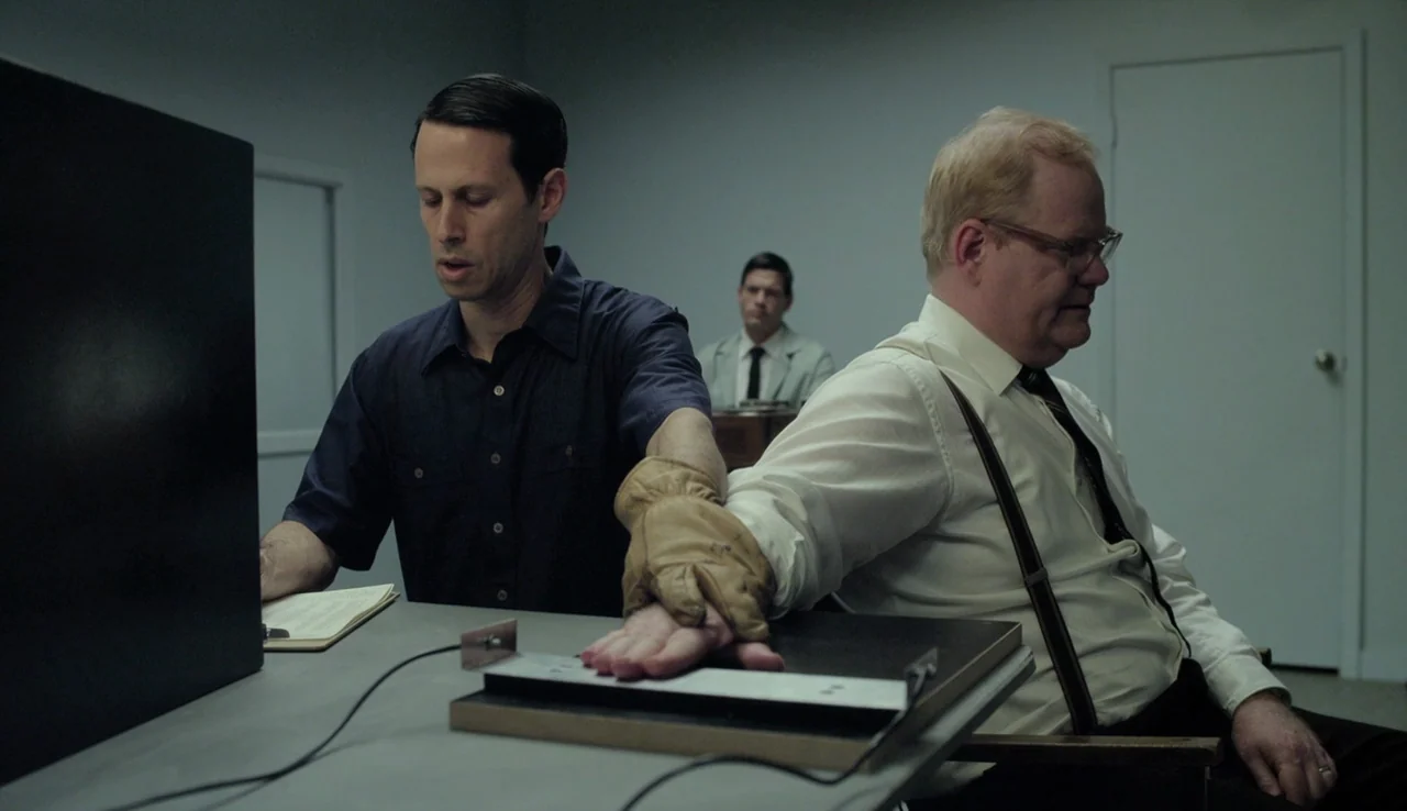 Experimenter (2015) movie still: medium — A spare, clinical room with grey walls and a plain door in the background. Two men sit at a metal desk…