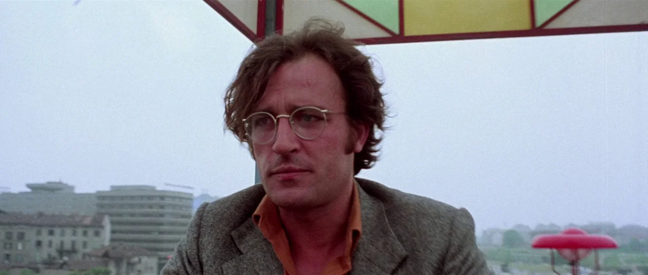 The Suspicious Death of a Minor (1975) movie still: closeup — A man stands on a rooftop with a blurred cityscape visible in the distance. He wears a…