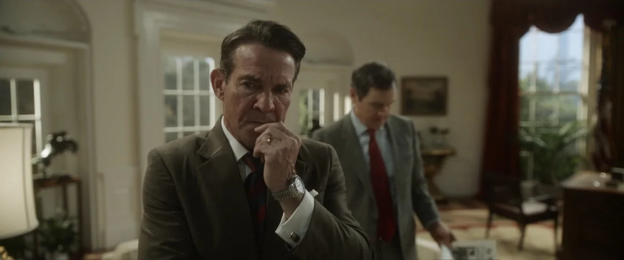 Reagan (2024) movie still: medium — A spacious formal interior with white molding and large windows draped in red fabric. A man in a suit stands in…