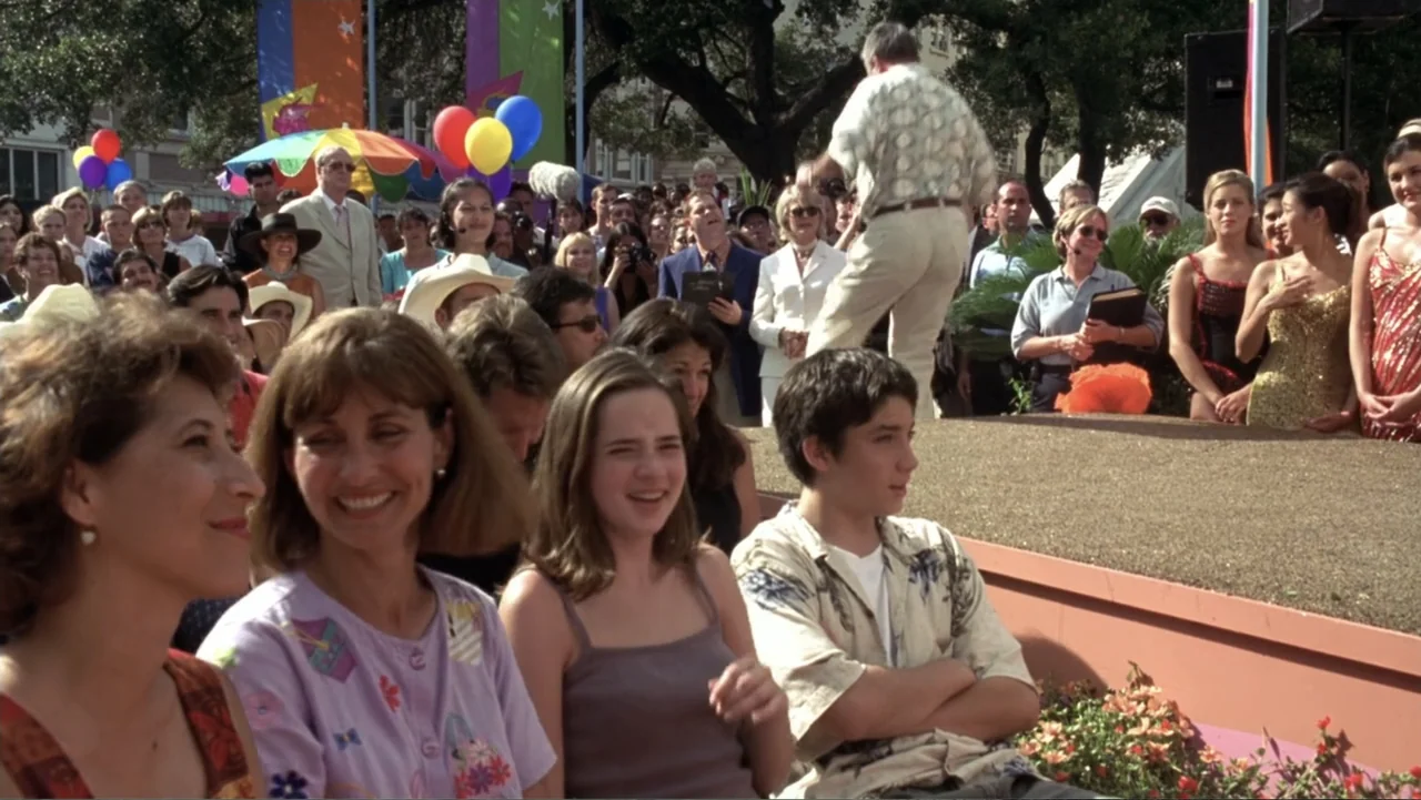 Miss Congeniality (2000) movie still: medium — A crowd gathers outdoors in a sunny area near a low stage platform. Men and women watch as an older…