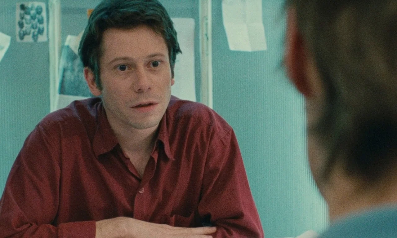 Late August, Early September (1998) movie still: medium — A man sits at a desk in a room with light blue walls. The background features papers…