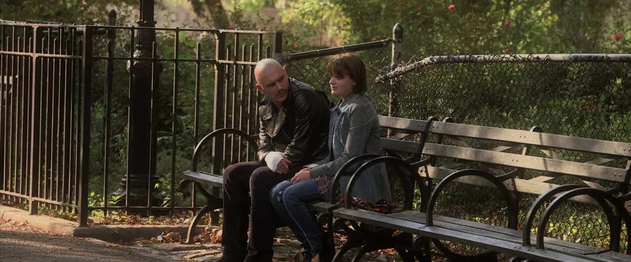 Zeroville (2019) movie still: fullbody — A wooden park bench sits next to a black metal fence in a green park. A man with a bandaged hand sits…