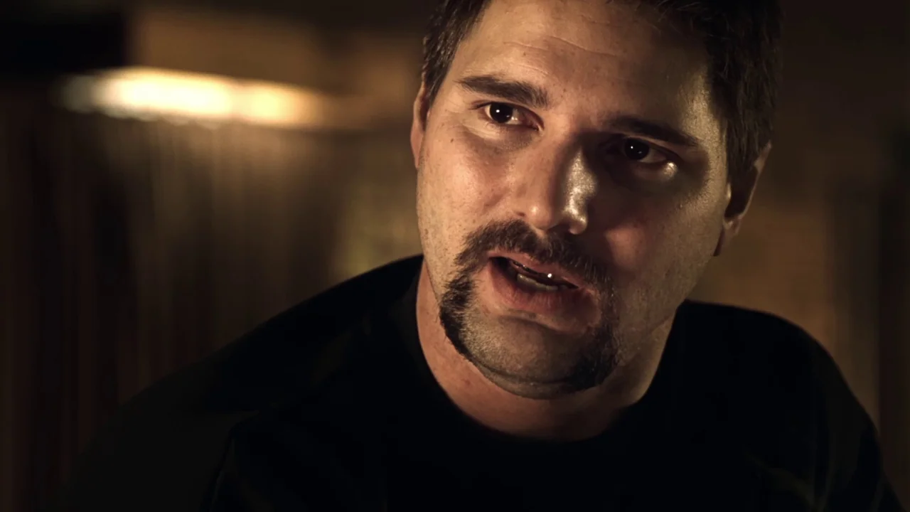 Chopper (2000) movie still: closeup — A man stands in a dimly lit indoor space with shadows obscuring the background. The man gazes forward with his…