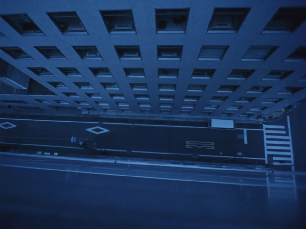 Tetsuo II: Body Hammer (1992) movie still: establishing — A top-down view shows an urban street flanked by a large office building. The asphalt road…