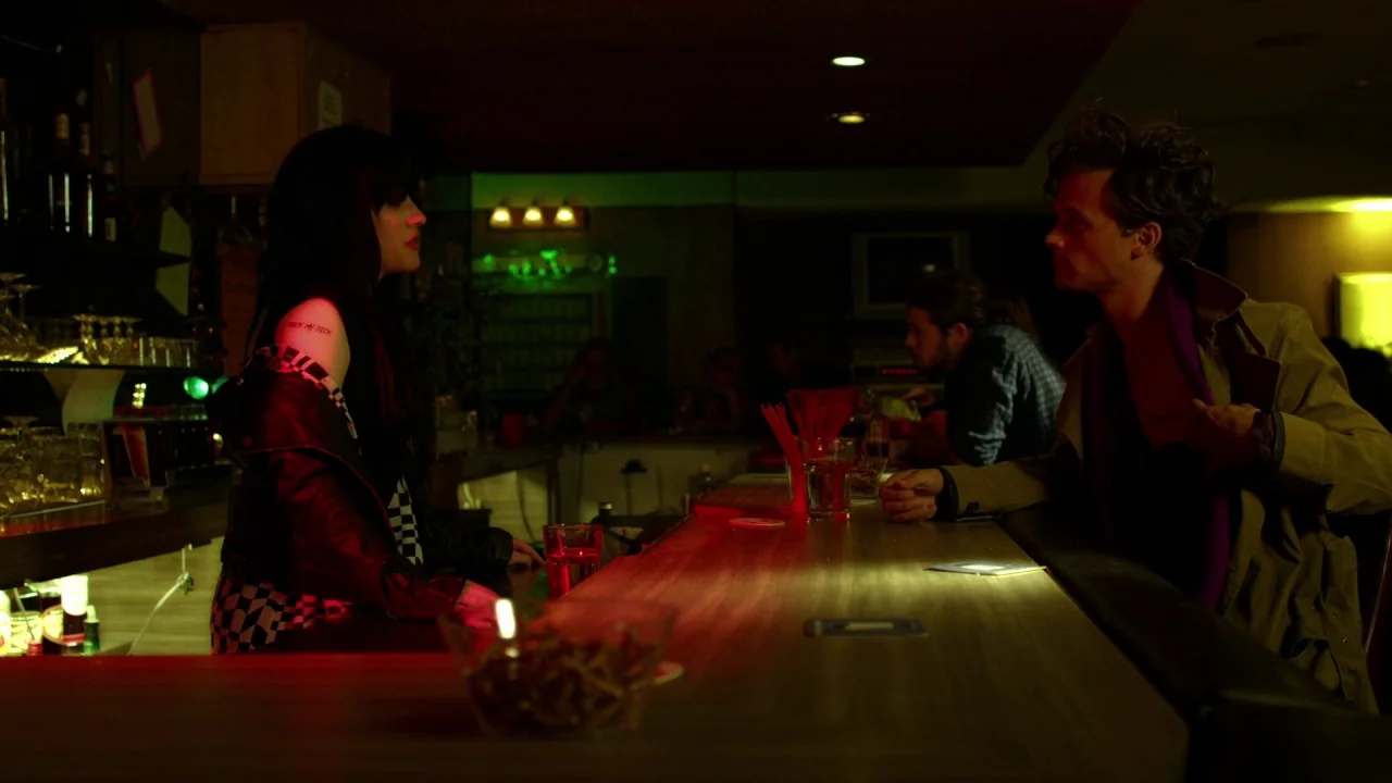 Suburban Gothic (2014) movie still: medium — A dimly lit bar features a long wooden counter under intense red and green lights. A woman in a leather…