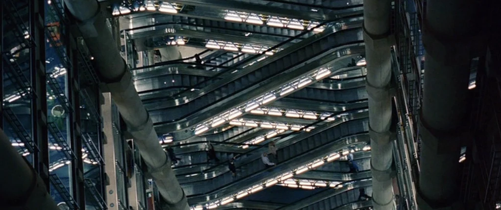 Code 46 movie still: establishing — A complex multi-level building interior features crisscrossing escalators and large cylindrical structural…