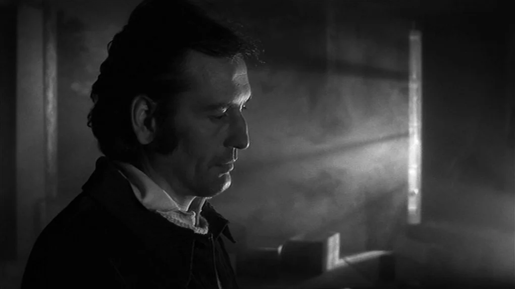 The State of Things (1982) movie still: medium — A man stands inside a dark, smoke-filled industrial room with light rays visible. The man looks…