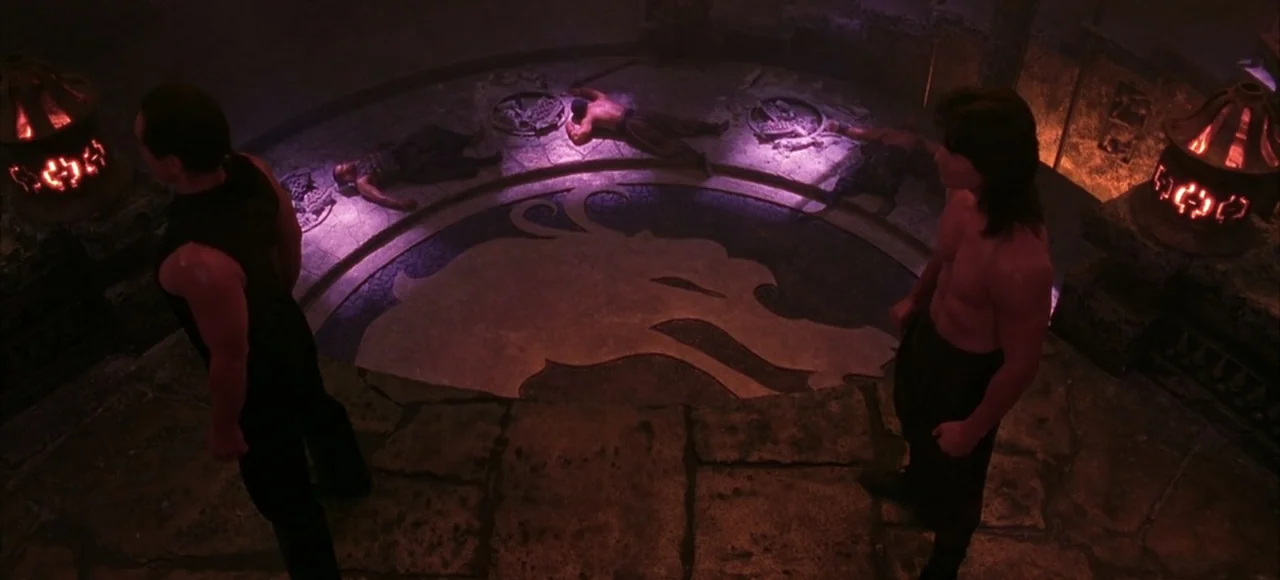 Mortal Kombat (2021) movie still: wide — An ancient stone circular floor features a dragon inlay within a dark chamber. Two men face each other with…
