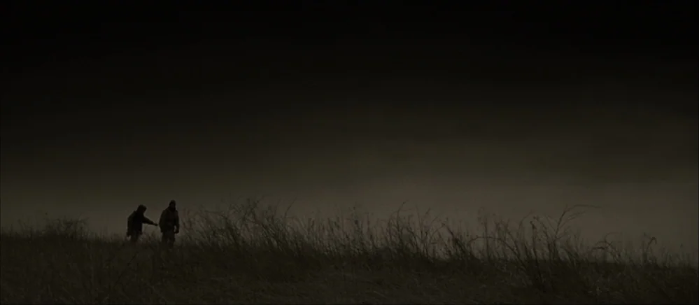 The Road (2009) movie still: wide — A vast, dark field under a heavy, overcast sky during the night. Two hooded figures walk across the dry,…