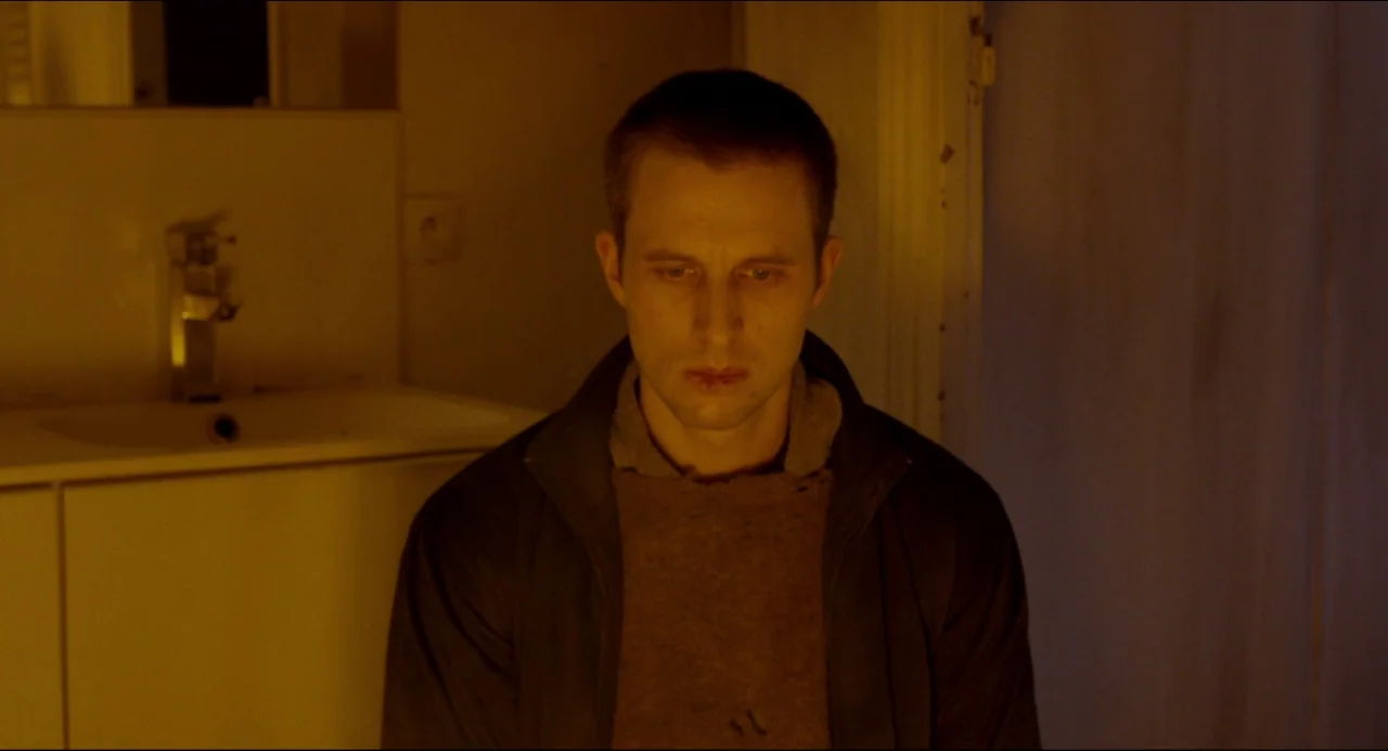 The Night Eats the World (2018) movie still: medium — A dark, cramped bathroom interior with warm, low-level illumination. A young man sits facing…